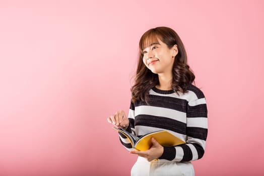 Portrait of beautiful Asian young woman teenage smiling holding a yellow book and reading, studio shot isolated on pink background with copy space