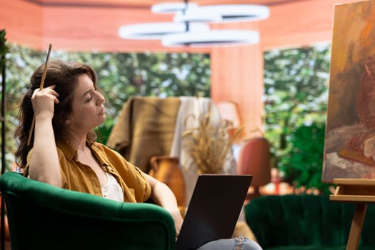 Female artist searching for a new painting inspiration on laptop, sitting in art and craft atelier and looking for creativity or imagination to create next masterpiece. Woman browses internet.