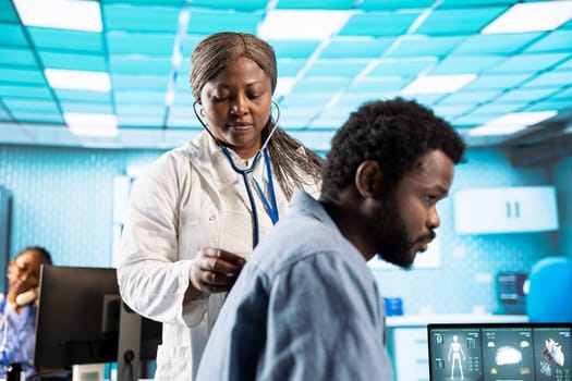African american doctor does a respiratory examination checkup with stethoscope, listening to heartbeat and lungs functioning normally. Medic specialist checking pulse for young man.
