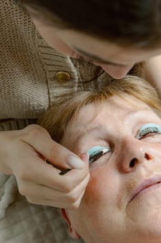 One Caucasian young beautiful brunette cosmetologist applies an adhesive with a sponge stick to the eyelashes of the right eye with a sponge stick of an elderly female client preparing them for the lamination procedure, which is lying on the cosmetology table in a home beauty salon, top side close-up view.
