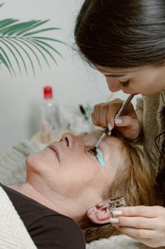 One Caucasian young beautiful brunette cosmetologist applies brown paint with a brush to the eyelashes of the left eye of an elderly woman client who is lying on the cosmetology table in a home beauty salon, side view close-up.