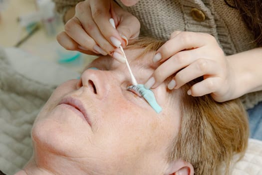 One Caucasian young unrecognizable girl cosmetologist applies a strengthening and healing agent with a cotton stick to the eyelashes of the left eye of an elderly female client who is lying on a cosmetology table in a home beauty salon, side view very close-up.