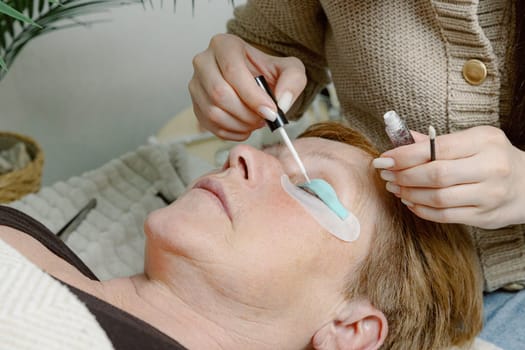 One Caucasian young unrecognizable girl cosmetologist applies glue to a silicone stand for the eyelashes of the left eye using a brush to an elderly female client who is lying on a cosmetology bed in a home beauty salon, side view close-up.