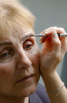 One young Caucasian young unrecognizable girl cosmetologist carefully paints the left eyebrow with brown paint using a brush to a beautiful elderly woman with gray-blue eyes looking to the side in a home beauty salon, side view close-up.