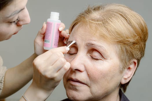 One young Caucasian young beautiful girl cosmetologist carefully removes with a cotton stick the remaining paint from the right eyebrow of an elderly woman with closed eyes, looking to the side using a cleanser, in a home beauty salon, close-up side view.