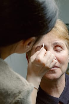 One young Caucasian young brunette girl cosmetologist carefully removes with a cotton stick the remains of paint from the right eyebrow of an elderly woman with closed eyes, looking to the side using a cleanser, in a home beauty salon, close-up side view.