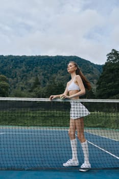 Energetic young female tennis player in a stylish white outfit exudes confidence on the court, set against a lush green mountainous background, ideal for sports promotion