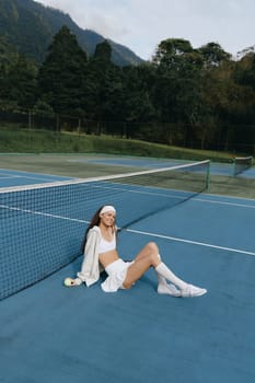 A cheerful young woman wearing sports clothing sits on a tennis court, surrounded by greenery Her joy and sporty style capture the essence of active living and fitness