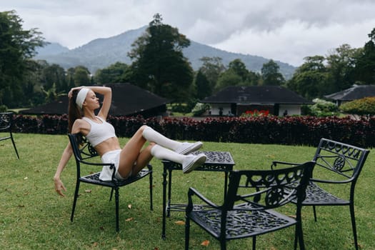 A fashion forward model dressed in a sporty ensemble, exuding relaxation and confidence while lounging on a chair amidst lush green hills and cloudy skies