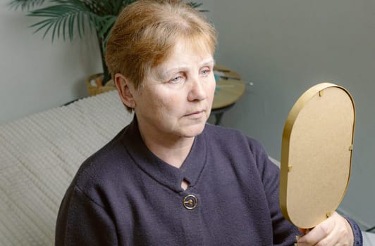 One young Caucasian elderly woman looks in the mirror, carefully examining the drawn white outline on her eyebrows before starting coloring while sitting in a home beauty salon on a cosmetology bed, top side view close-up.