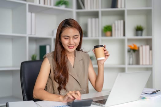 A focused young professional sits in her modern office workspace, sipping coffee and calculating important figures on her calculator, showcasing a productive work atmosphere.