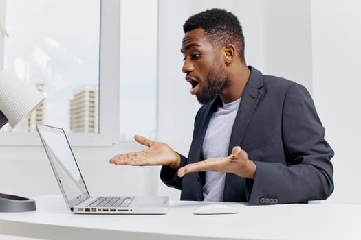 Stressed businessman in formal attire holding head in frustration while working on laptop at cluttered office desk.