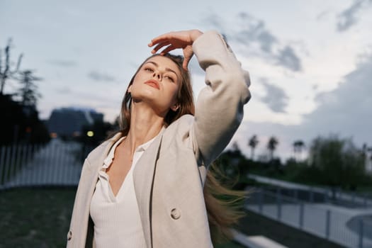 Confident woman wearing beige coat and white top poses outdoors at sunset, touching hair with relaxed expression, soft lighting, casual fashion style, urban environment