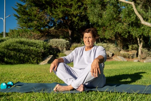 Fit senior woman in activewear sitting on yoga mat in park on a sunny summer day, enjoying a healthy and active lifestyle outdoors