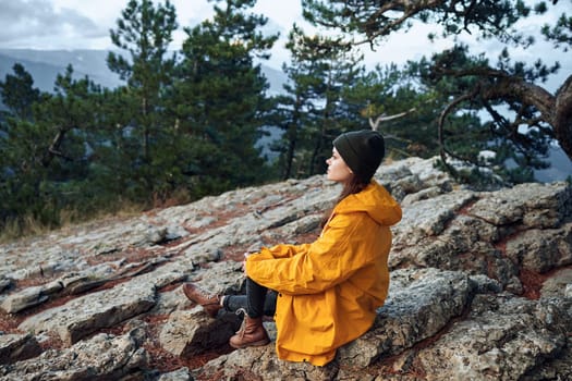 Woman traveler in a vibrant yellow raincoat sitting peacefully on a rock in front of a serene tree