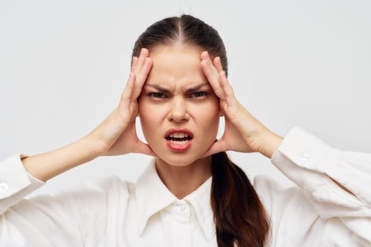 a woman in a white shirt is visibly frustrated, pressing her hands against her temples in a light gray background The expression captures stress and annoyance, depicting a relatable emotional state