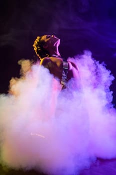 Sensual woman in silver underwear kneeling in cloud of fume and bending back under neon illumination while dancing in dark room