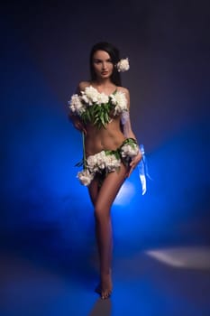Gorgeous young self assured female model with long dark hair and fresh white flowers covering naked body standing in dark studio - smoke under neon light