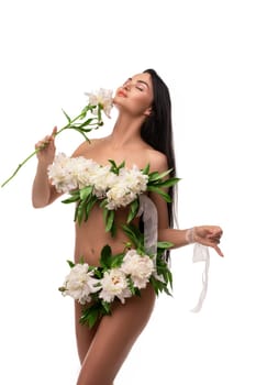 Self assured young naked female model with long dark hair covering body with bunch of fresh delicate flowers and looking at camera against white background