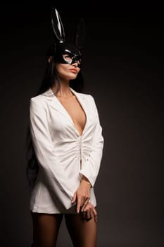 Confident young female in elegant white dress and black carnival mask standing and looking at camera isolated on black background