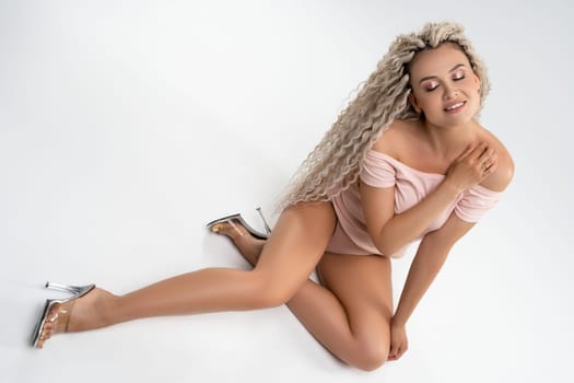 Sensual young ethnic female with curly hair wearing white lingerie and looking at camera while sitting on white background in studio