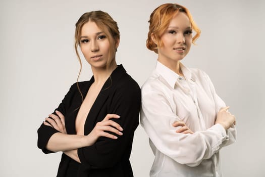 Positive confident young female colleagues in elegant outfits standing back to back with crossed arms and looking at camera against gray background