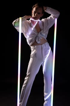 Confident young lgbt male model in white shirt and pants with makeup touching face and looking at camera while standing under neon lights