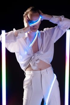 Confident young androgynous male model in white shirt and pants with makeup touching face and looking at camera - black studio near LED sticks