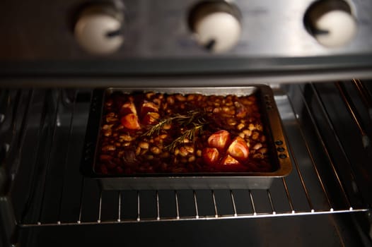 A baking tin filled with baked beans and tomatoes, flavored with rosemary, inside a preheated oven. The warm lighting enhances the inviting and appetizing nature of the dish during preparation.