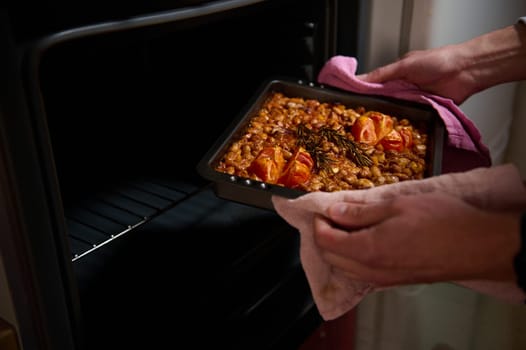 Carefully removing a baking tray of freshly cooked beans garnished with tomatoes and rosemary from the oven, evoking homely feelings of warmth and nourishment.