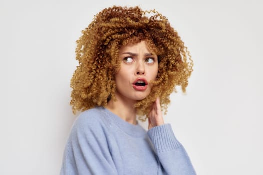A surprised young woman with voluminous curly hair, wearing a pastel blue sweater, poses against a light neutral background, highlighting her emotional expression and unique hairstyle