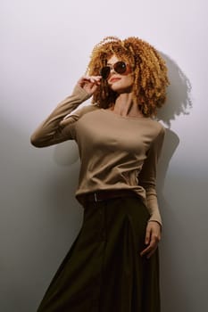 A fashionable woman posing confidently against a gray background Her curly hair frames her face, and she wears stylish sunglasses, showcasing a blend of elegance and contemporary style