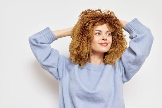 A smiling woman with curly hair poses playfully in a light blue sweater against a minimalistic white background Her joyful expression and relaxed pose convey a sense of natural beauty and confidence