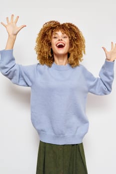 Cheerful woman with voluminous curly hair wearing a cozy blue sweater, arms raised in excitement The light background enhances her lively expression, perfect for themes of happiness and celebration