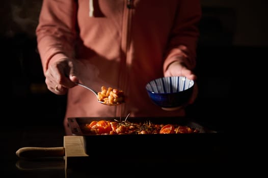 A person serves freshly cooked baked beans garnished with herbs into a blue bowl, captured in a dimly-lit kitchen setting emphasizing deliciousness and care in meal preparation.