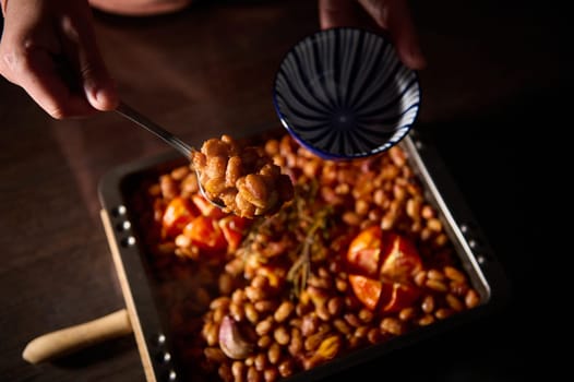 Hands serve a portion of baked beans, roasted tomatoes, and garlic into a patterned ceramic bowl, emphasizing the dish's rustic deliciousness.