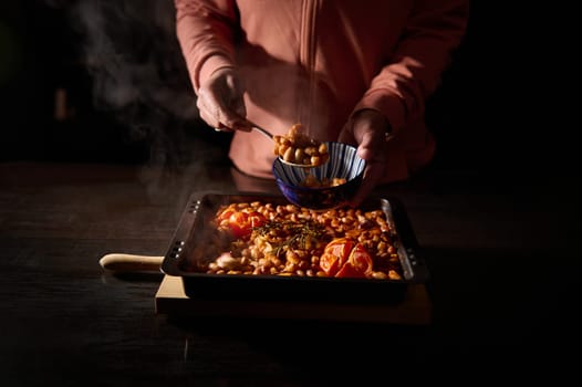 Person serving a hearty tray of baked beans and tomatoes into a bowl, set under warm, rustic lighting. A steaming tray of comforting food serves a cozy meal in an intimate setting.