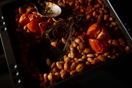 A rustic presentation of baked beans topped with roasted tomatoes and rosemary sprigs on a dark-toned baking tray, showcasing natural cooking and hearty flavors.