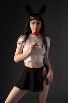 Confident young female model in stylish outfit and bunny ears looking at camera while standing against black background and lick candy