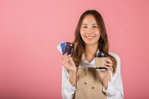 Portrait Asian happy young woman barista bar tender coffee maker holding coffee tea hot cup and credit card studio shot isolated on pink background, female smiling hold takeaways cup for payment
