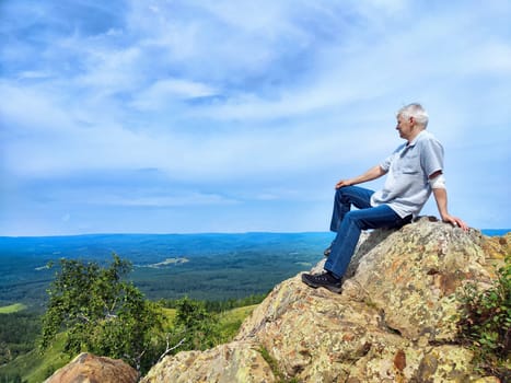 An elderly man sits on a large rock, gazing over a vast landscape of rolling hills and forests under a clear blue sky.