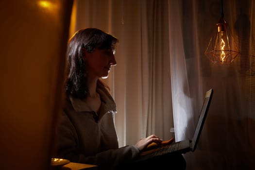 In a homely apartment, a middle-aged woman finds relaxation as she works on her laptop, illuminated by soft evening light.