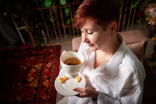 A mature woman with short hair sips tea and smiles while seated comfortably in a cozy, decorated space.