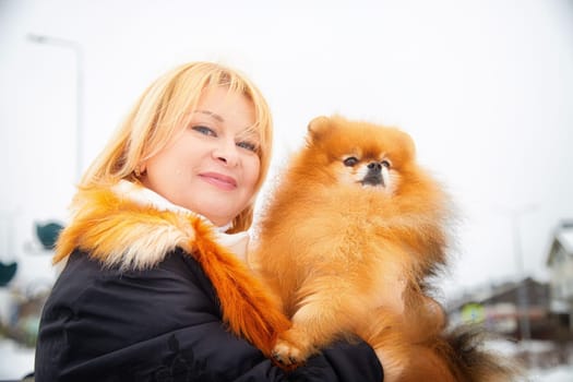 Blonde woman holds her adorable Pomeranian as they stroll through a snowy landscape, radiating warmth and joy.
