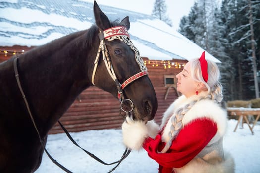 A beautiful girl in a national dress interacts affectionately with a horse during the Maslenitsa celebration in winter.