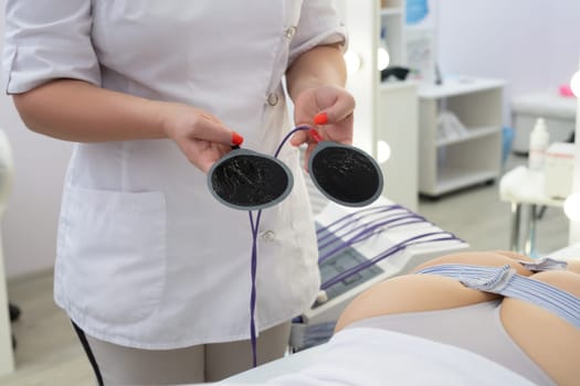 Crop unrecognizable masseuse in white medical robe attaching electronic anti cellulite pads on buttocks of female client lying on bed in modern clinic