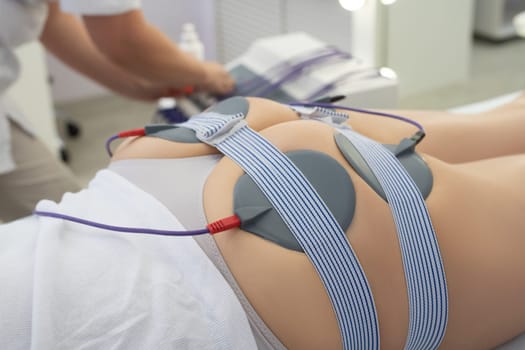 High angle of crop anonymous client with anti cellulite electronic pads on buttocks lying on couch during lymphatic drainage treatment with doctor