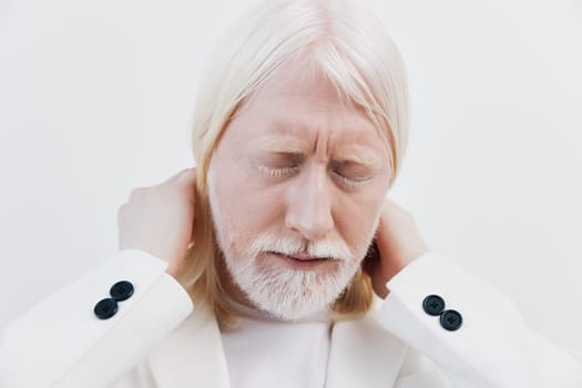 An elderly man with long white hair and a white beard dressed in a white suit shows a pained expression while holding his neck The background is a soft, neutral white, emphasizing his discomfort