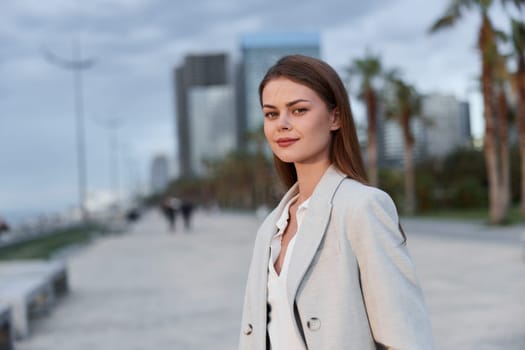 A confident woman in a fashionable blazer stands by the seaside, showcasing a modern business aesthetic. The scene captures her poise and determination against a contemporary urban backdrop.