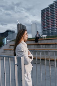 Thoughtful woman wearing a neutral coat stands by a metal fence in a cityscape with modern buildings and overcast sky, reflecting introspection and contemporary fashion style